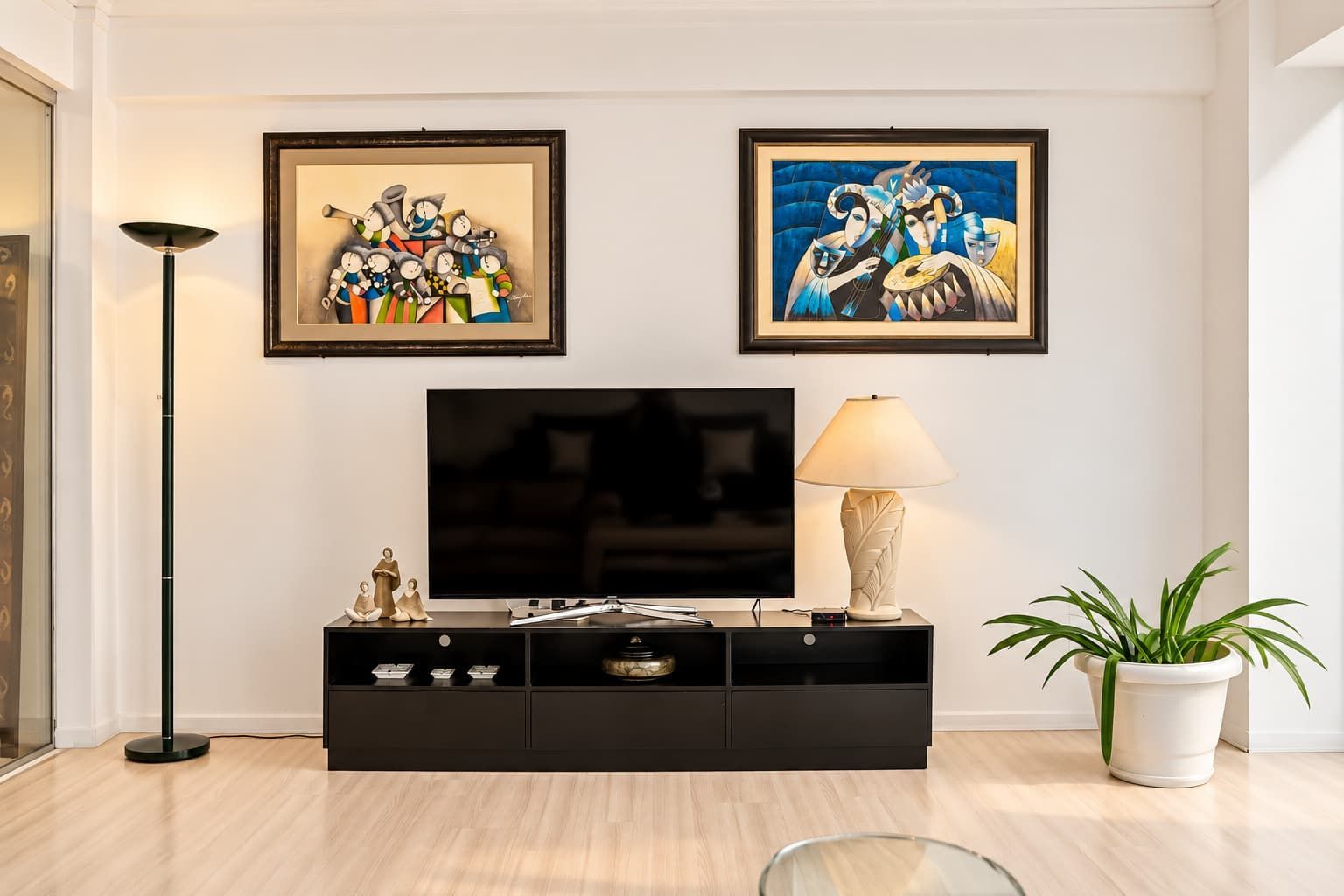 A modern living room with a flat-screen TV, a black entertainment unit, a floor lamp, and two framed artworks on the wall.