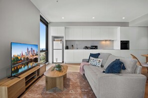 Ein modernes Wohnzimmer mit einem Flachbildfernseher, einem grauen Sofa, einem Holztisch und Blick auf die Stadtlandschaft.