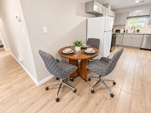Ett modernt kök med vita skåp, ett kylskåp i rostfritt stål och en matplats med ett runt träbord och mönstrade stolar.