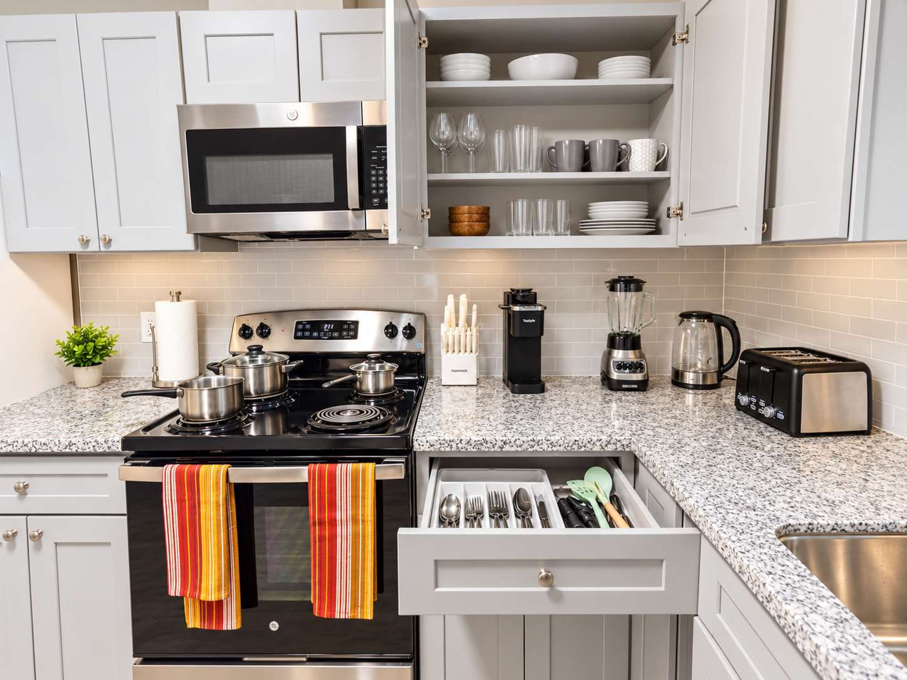 A modern kitchen with white cabinets, a stainless steel stove, a microwave, a toaster, a blender, and a coffee maker.