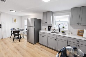 A modern kitchen with grey cabinets, a stainless steel refrigerator, a dining area with a table and chairs, and a window with blinds. - FIFA 2 Apt Bundle Near NRG  DT   Med Center - 12 (Houston)