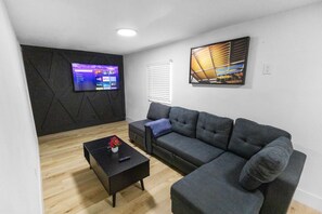Ein modernes Wohnzimmer mit einer Eckcouch, einem Couchtisch mit Fernbedienung, einem Flachbildfernseher an einer dunklen Wand und einem Gemälde über der Couch.