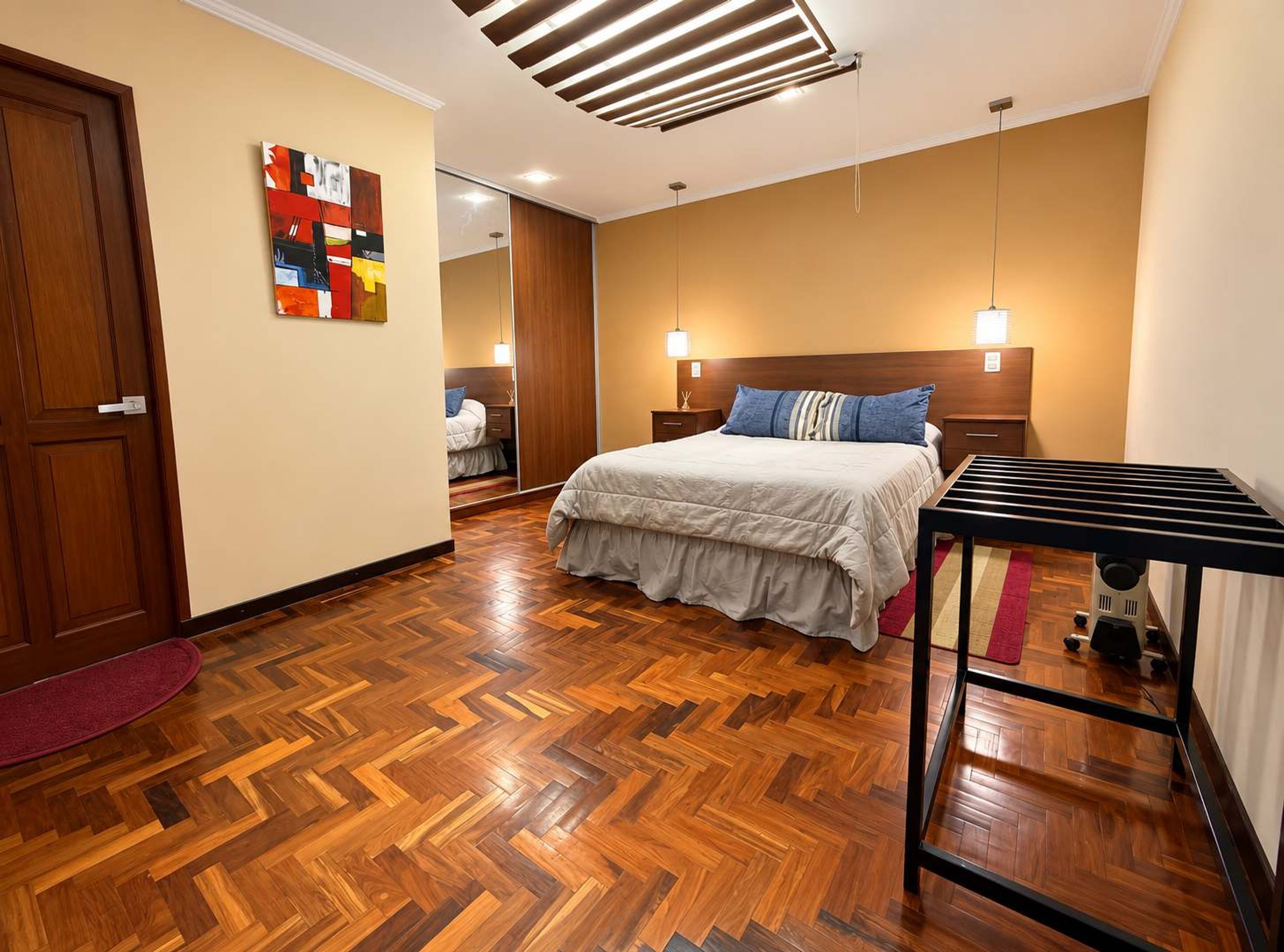 Une chambre avec un parquet en bois disposé en chevrons, un lit avec une couette blanche, une table de chevet, une commode, une lampe fixée au mur et une porte.