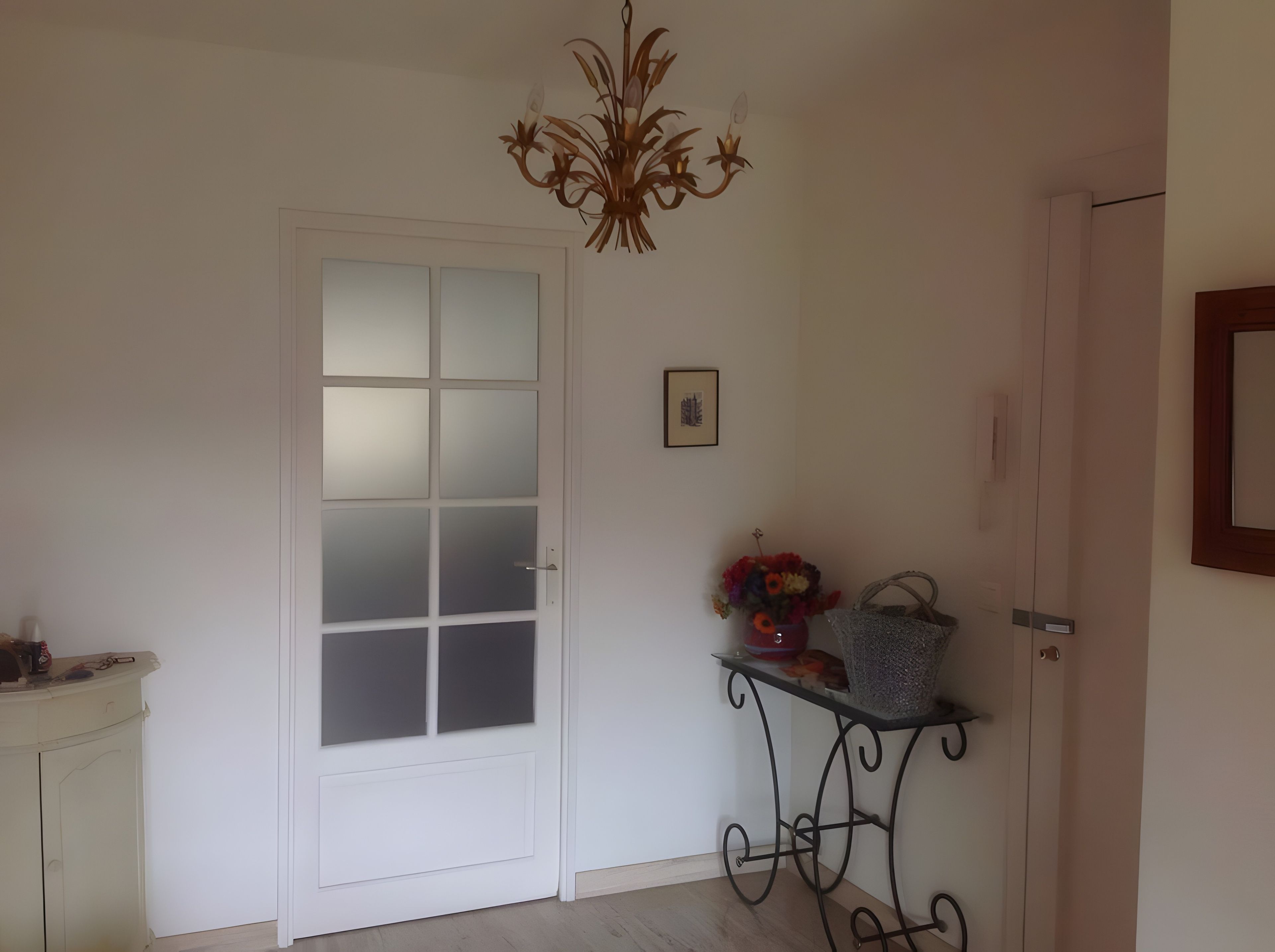 A room with a white door, a chandelier, a framed picture, and a small table with a basket.