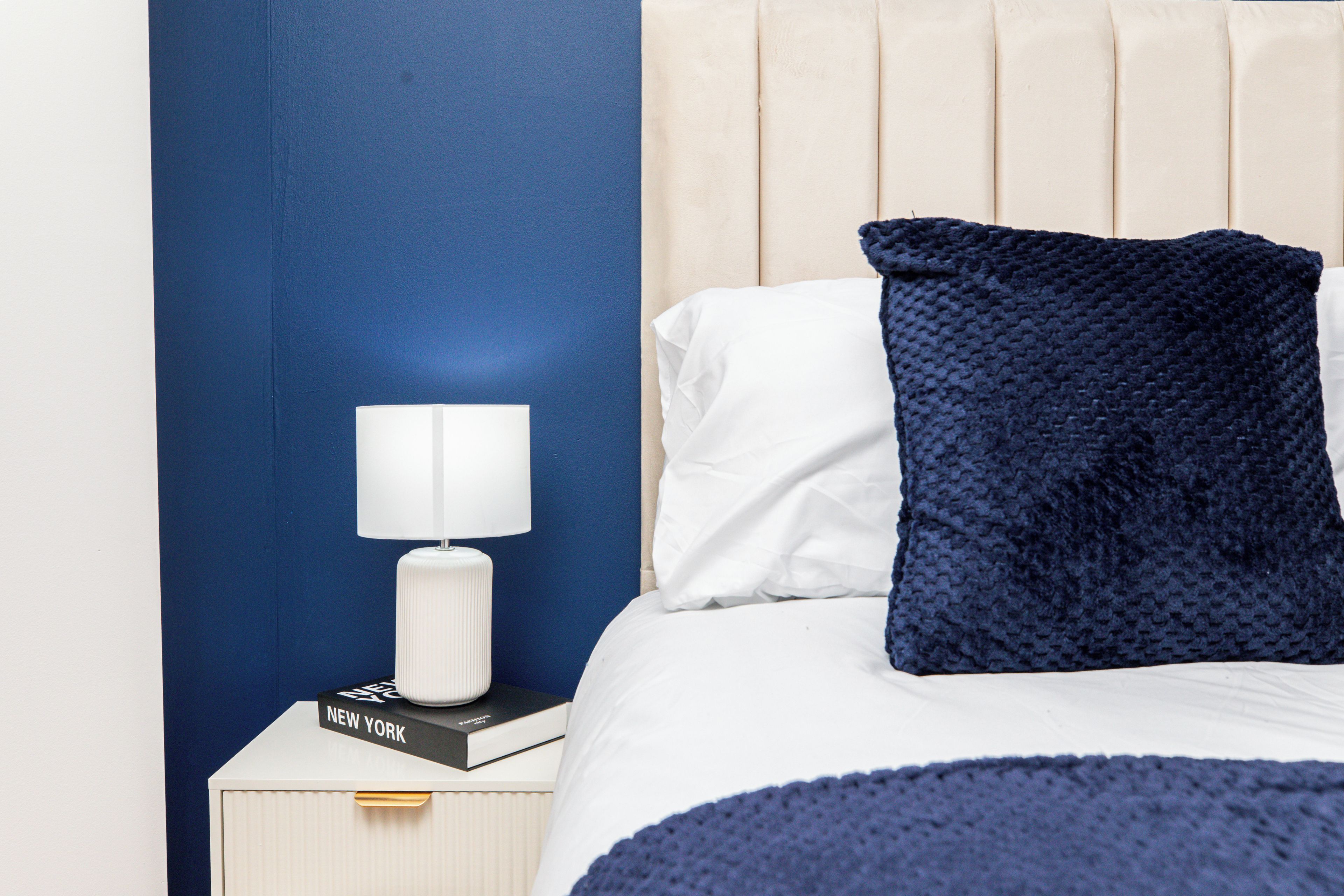 A neatly made bed with a white pillow and a dark blue textured pillow, a white bedside lamp, and a book titled 'NEW YORK' on a white nightstand.