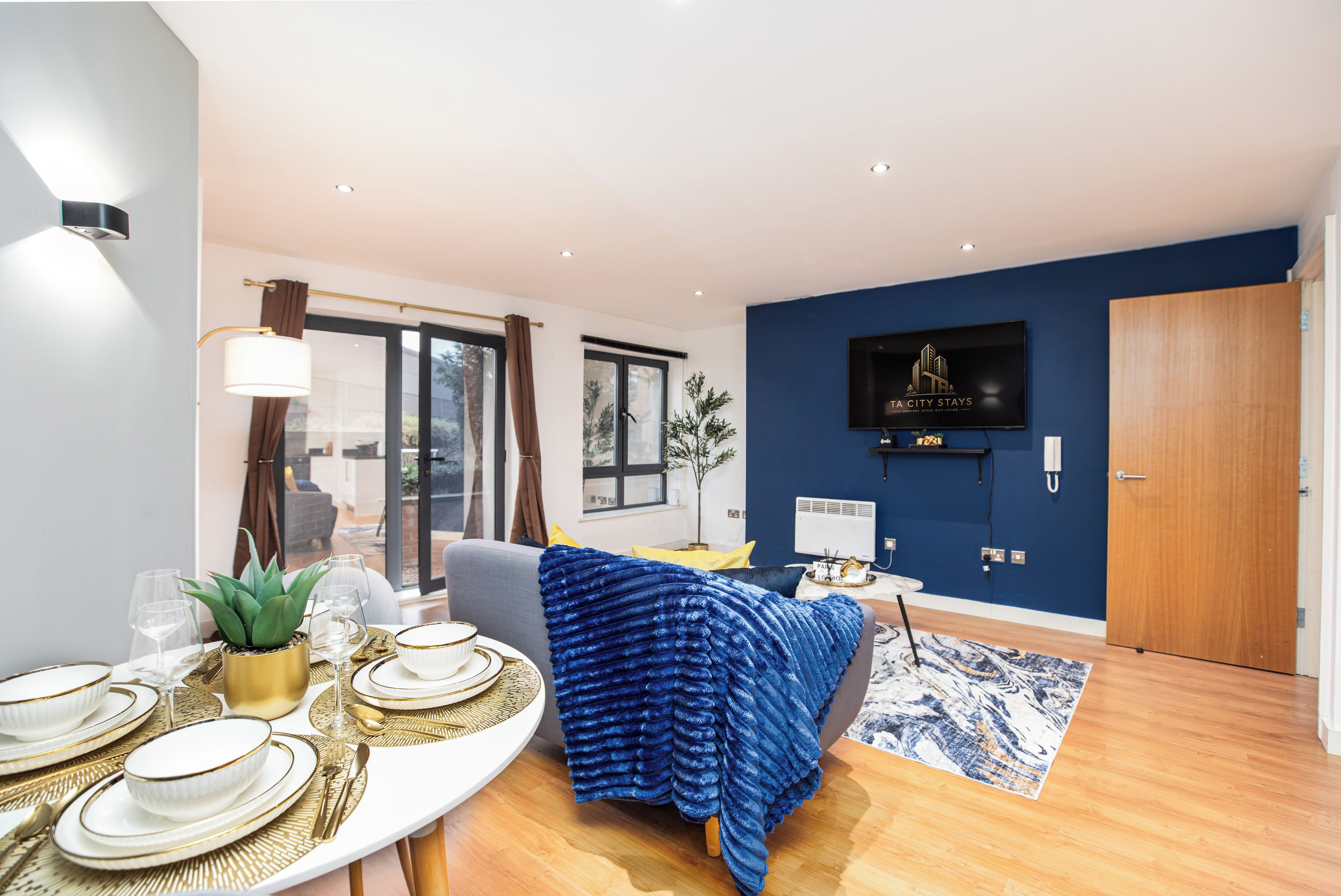 A modern living room with a dining area, a flat-screen TV mounted on the wall, and a large window.