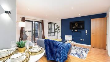 A modern living room with a dining area, a flat-screen TV mounted on the wall, and a large window.