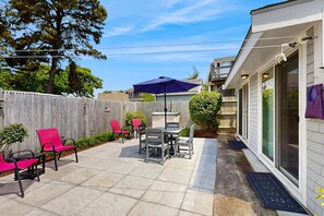 A patio with a table and chairs, a wooden fence, and a house with a balcony. - Cozy Cottage with Deck, Yard & Walk to the Beach (Dennis)