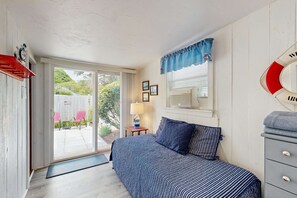 A bedroom with a bed, a window with a view, a bedside table, and a wall-mounted lamp. - Cozy Cottage with Deck, Yard & Walk to the Beach (Dennis)