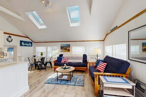 Sala de estar bem iluminada com sofá, mesa de centro e área de jantar com cadeiras. Há janelas com persianas, um relógio e uma placa que diz 'Beach'.