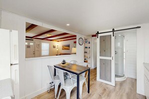 A compact kitchen with white cabinets, a wooden ceiling, and a dining area with a table and chairs. - Adorable Cottage With Patio & Walk to Sea St Beach (Dennis)