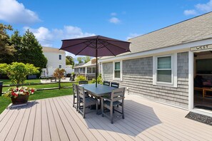 A deck with a table and chairs, a patio umbrella, and a house with a 'WEST' sign. - Adorable Cottage With Patio & Walk to Sea St Beach (Dennis)