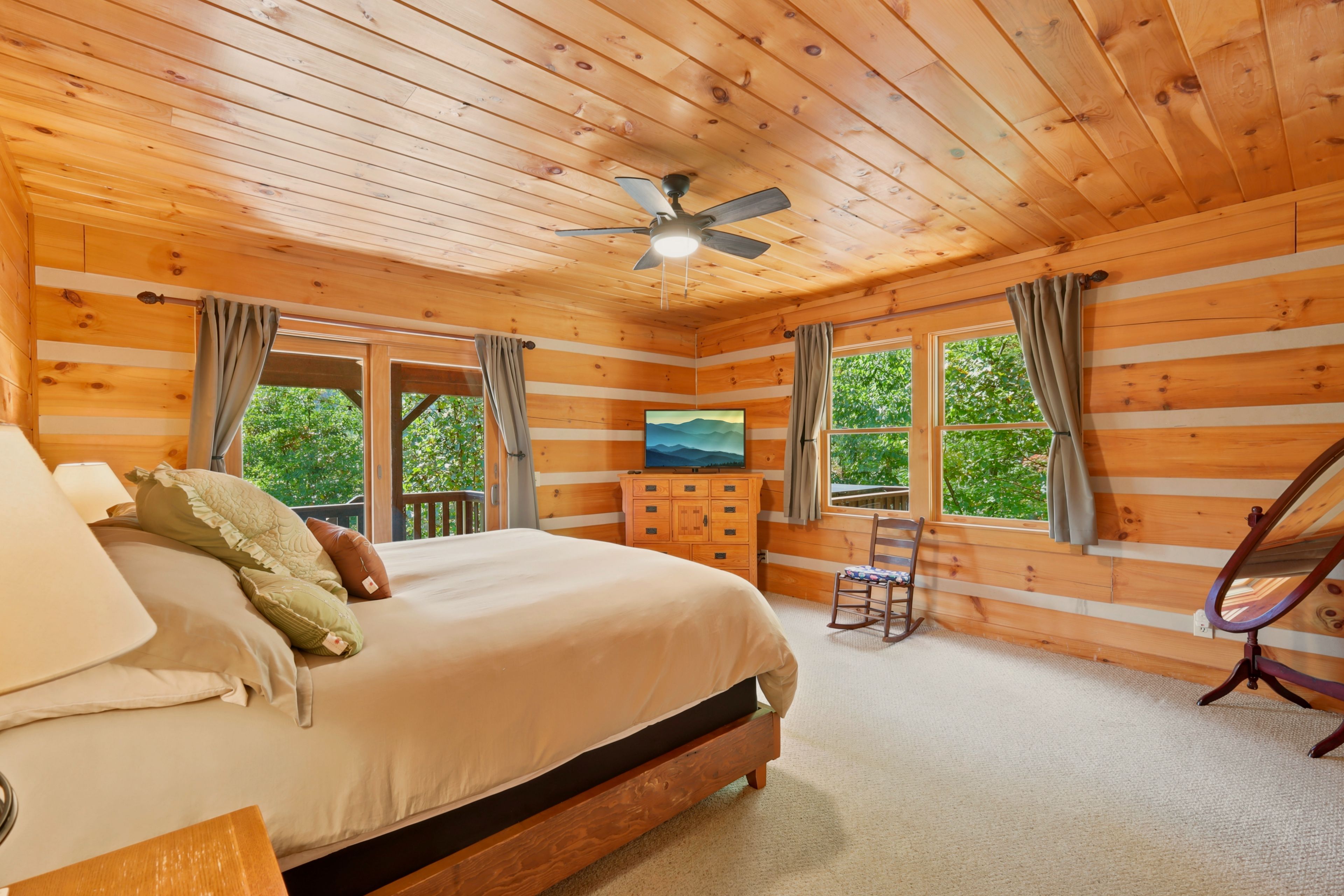 A wooden cabin bedroom with a bed, a TV, a chair, and a ceiling fan.