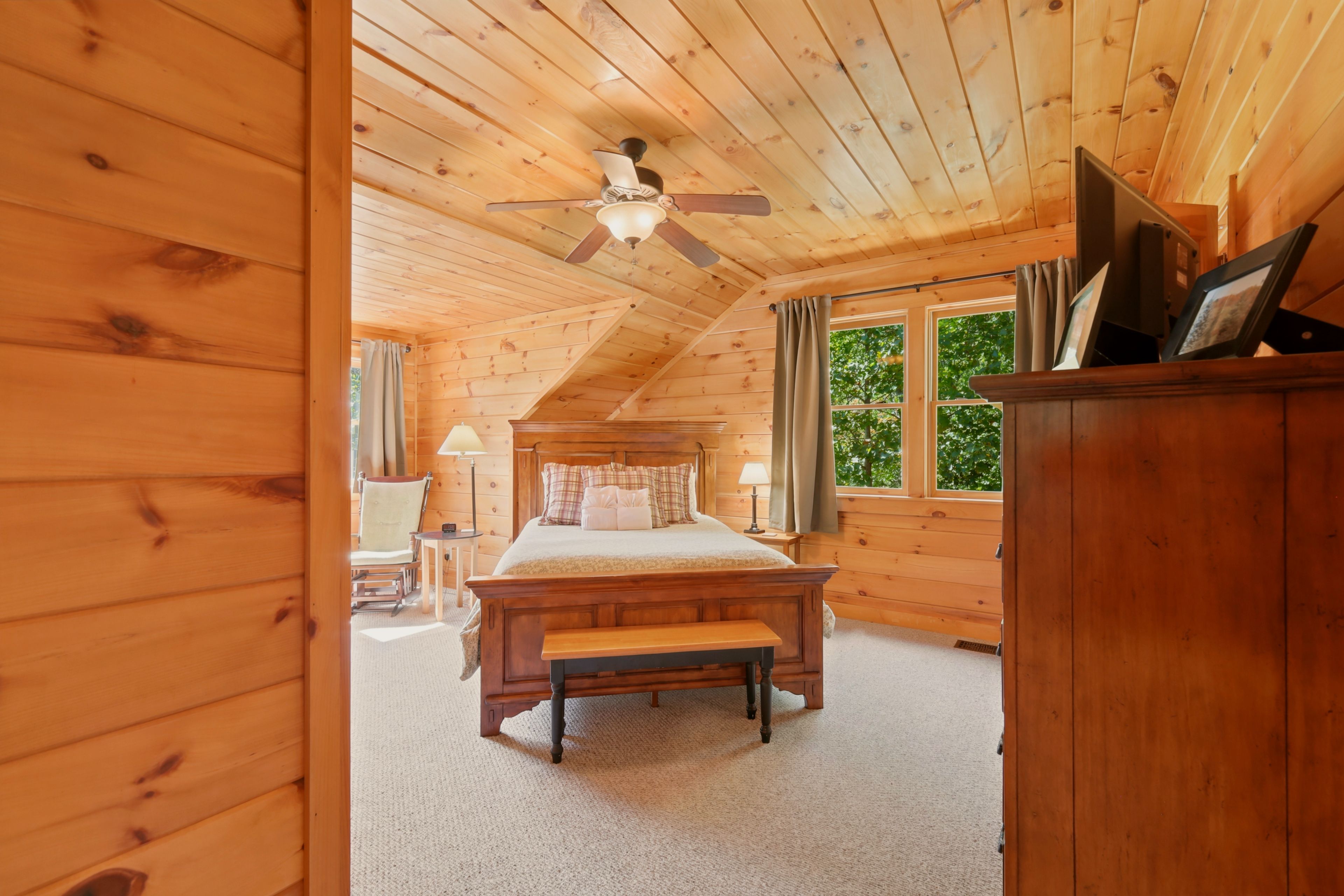 A wooden cabin bedroom with a bed, a bench, a ceiling fan, and a window with curtains.