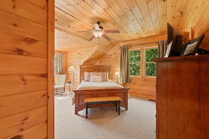 A wooden cabin bedroom with a bed, a bench, a ceiling fan, and a window with curtains.