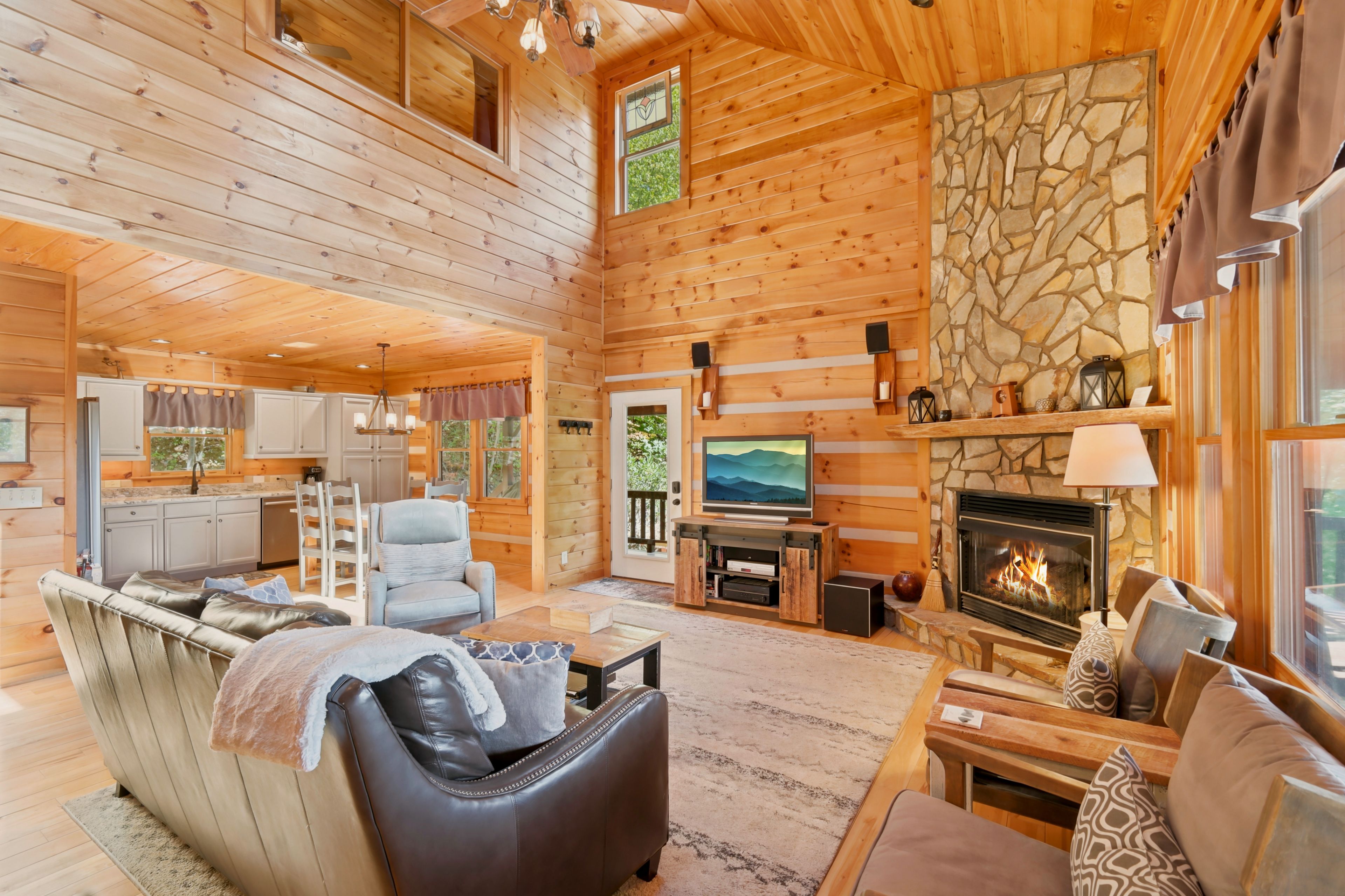 A log cabin interior with a fireplace, seating area, kitchen, and stone wall.