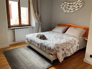 A bedroom with a bed, a radiator, a window with curtains, and a wall decoration. - BERGAMO ALTA SUITE - Spacious apartment in the heart of Bergamo Alta   (Bergamo)