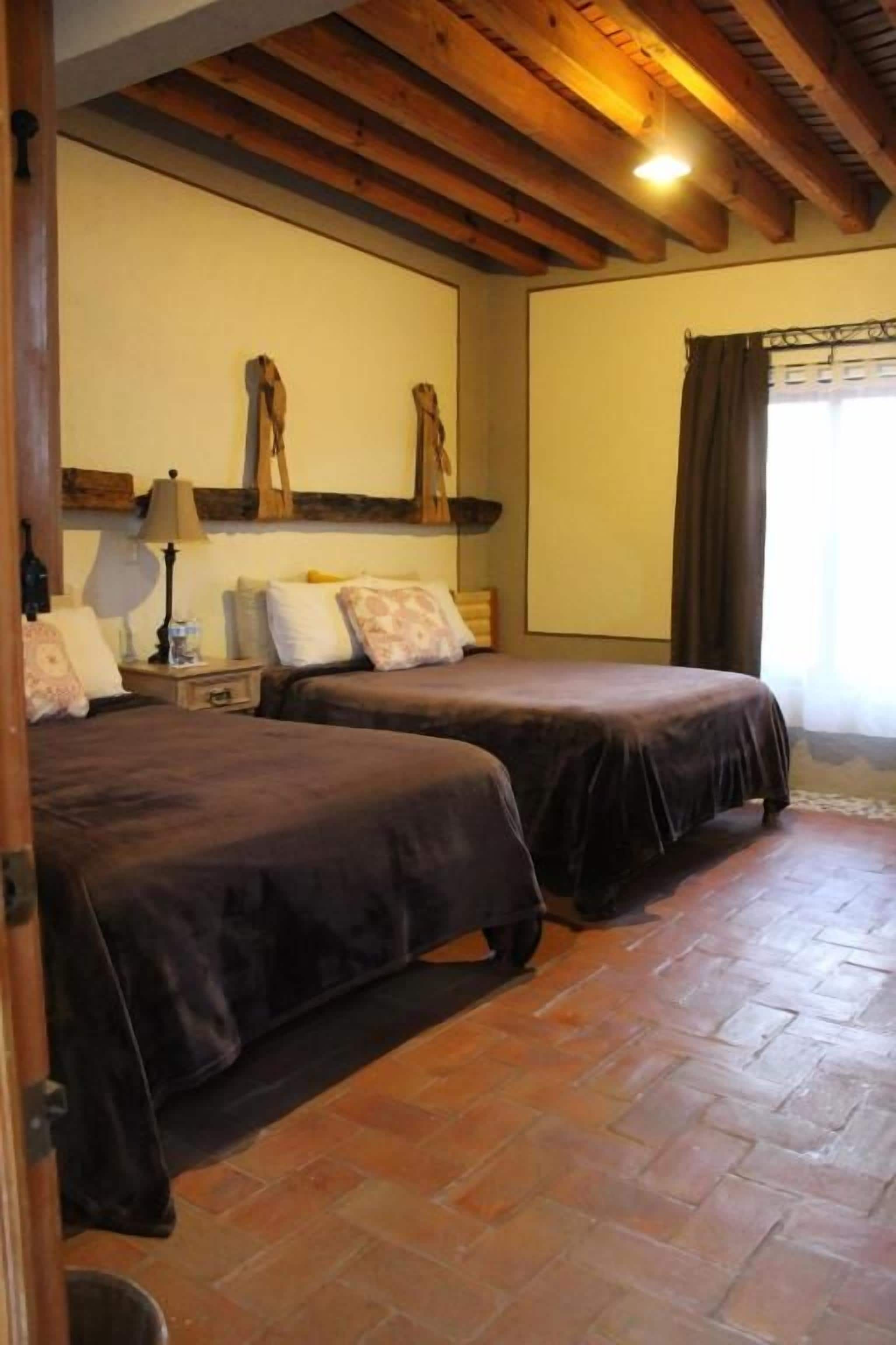 A room with two beds, a wooden ceiling, a brick floor, and a window with curtains.