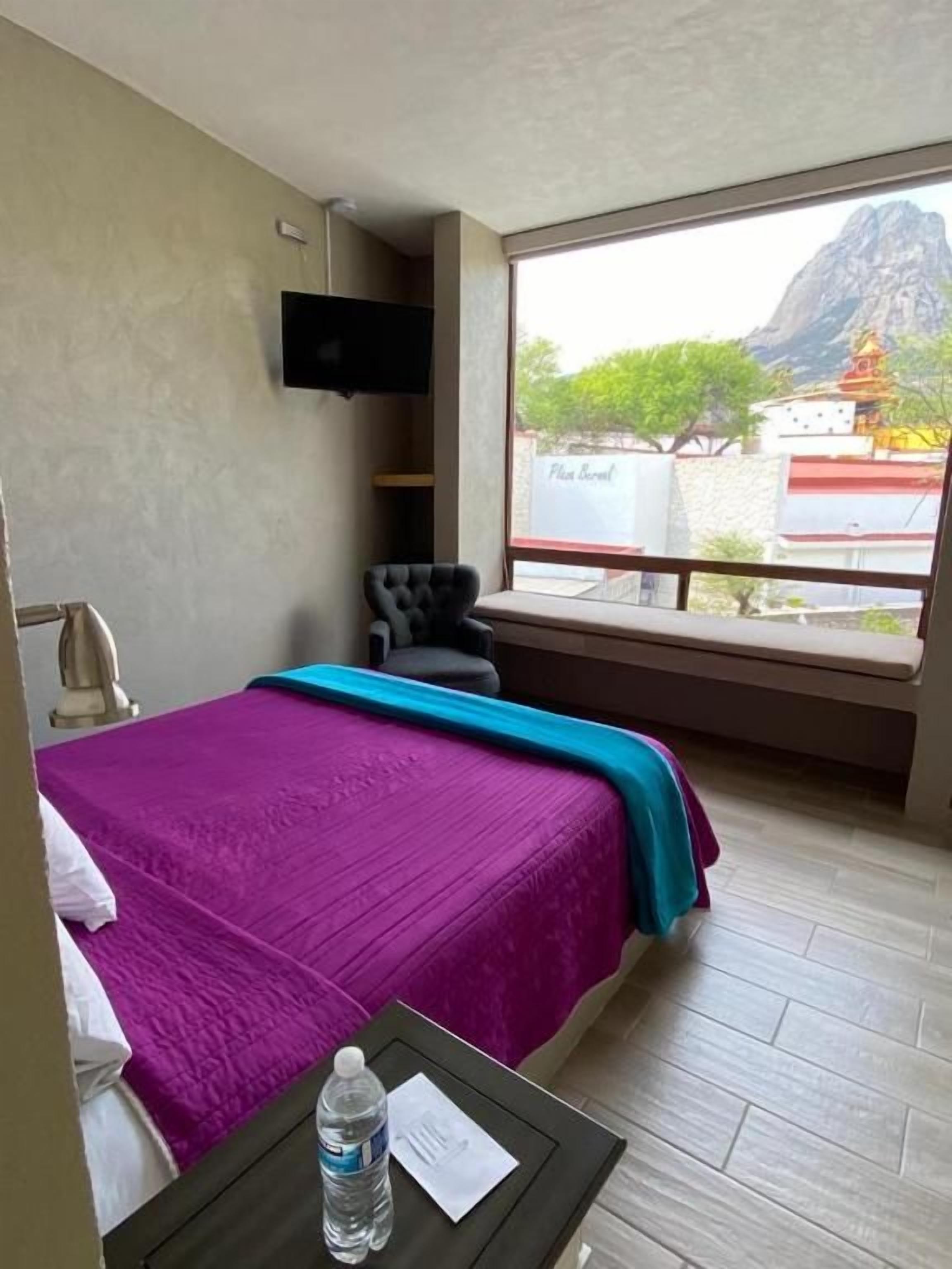A hotel room with a bed, a small table with a bottle, a chair, a TV, and a window with a view of a mountain and a temple.