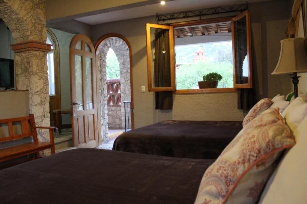 A room with two beds, a wooden bench, a stone pillar, and a window with a view of greenery.
