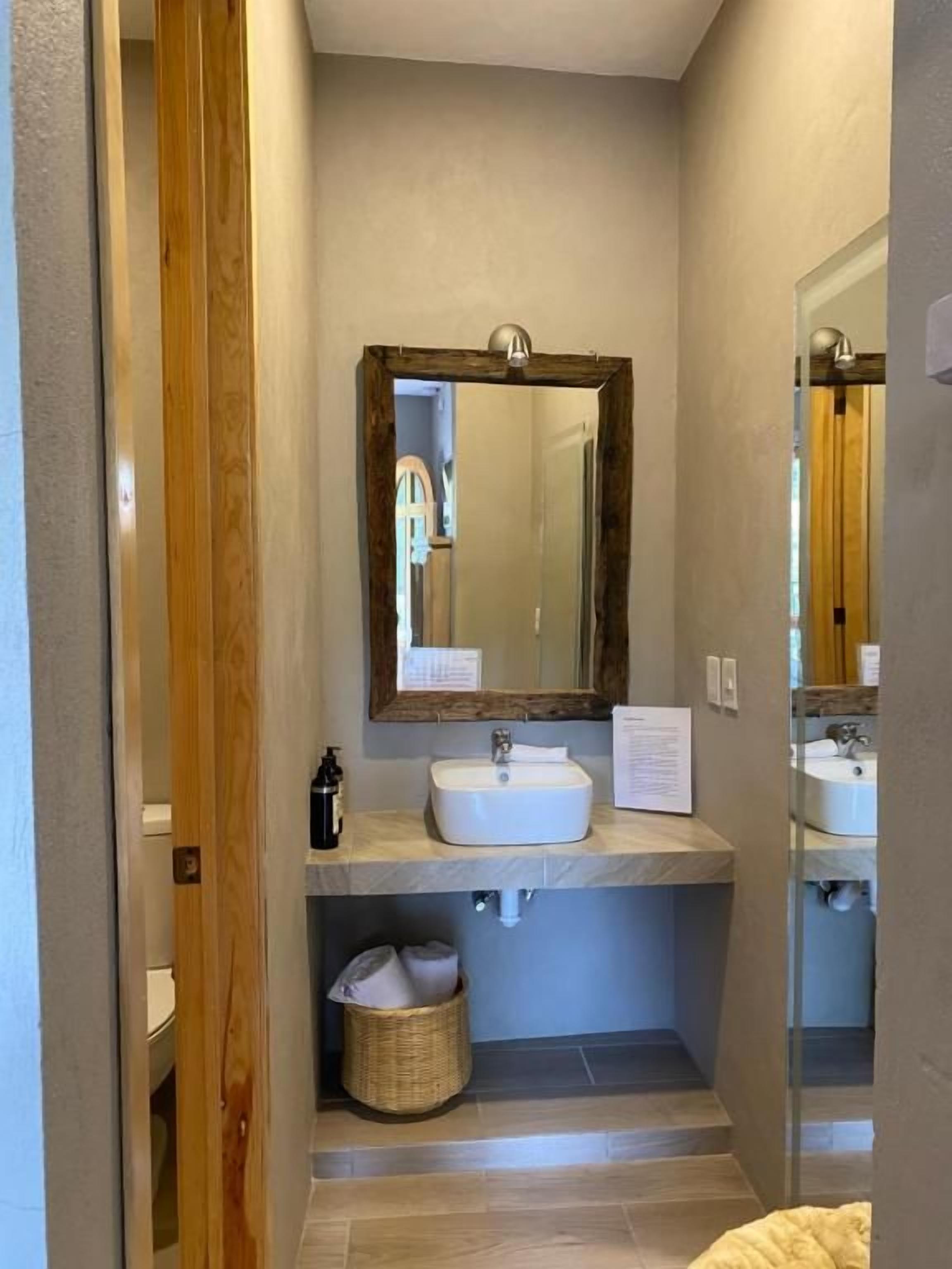 Un baño con puerta de madera, un lavamanos blanco, un espejo y una cesta.