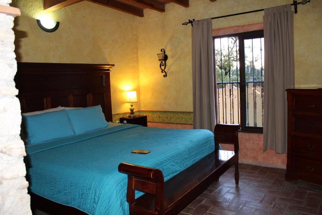 A bedroom with a bed, a nightstand, a lamp, a window with curtains, and a view of the outside.