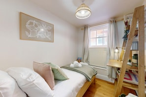 Une chambre à coucher avec un lit, des oreillers, une table de chevet avec une lampe, une fenêtre avec des rideaux et une œuvre d’art accrochée au mur.
