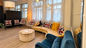 Ein modernes Wohnzimmer mit einer gebogenen Couch, einem runden Hocker und einem Flachbildfernseher. Mehrere Sessel mit Kissen im britischen Stil, und das Zimmer verfügt über große Fenster mit Tageslicht.