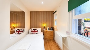 A bedroom with a bed, bedside lamps, a nightstand, a dresser, a mirror, and a window with a view of a building. - East London Compact Bedroom, Sleeps 1 With Wifi (London)