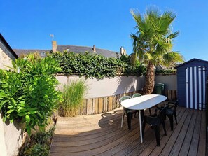 A wooden deck with a table and chairs, surrounded by greenery and a blue shed. - Charming house in the heart of the salt marshes (Guérande)
