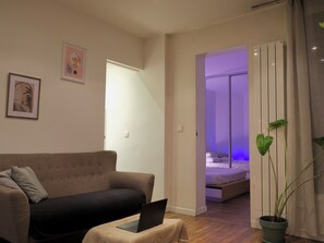 Ein Wohnzimmer mit einer Couch, einem Laptop auf einem kleinen Tisch, einer Topfpflanze und einem Bett im Hintergrund.