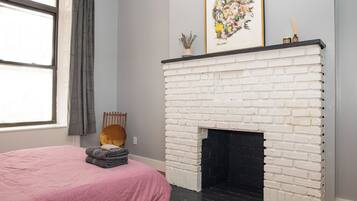 A room with a white brick fireplace, a bed with a pink bedspread, a window with curtains, and a framed artwork above the fireplace.