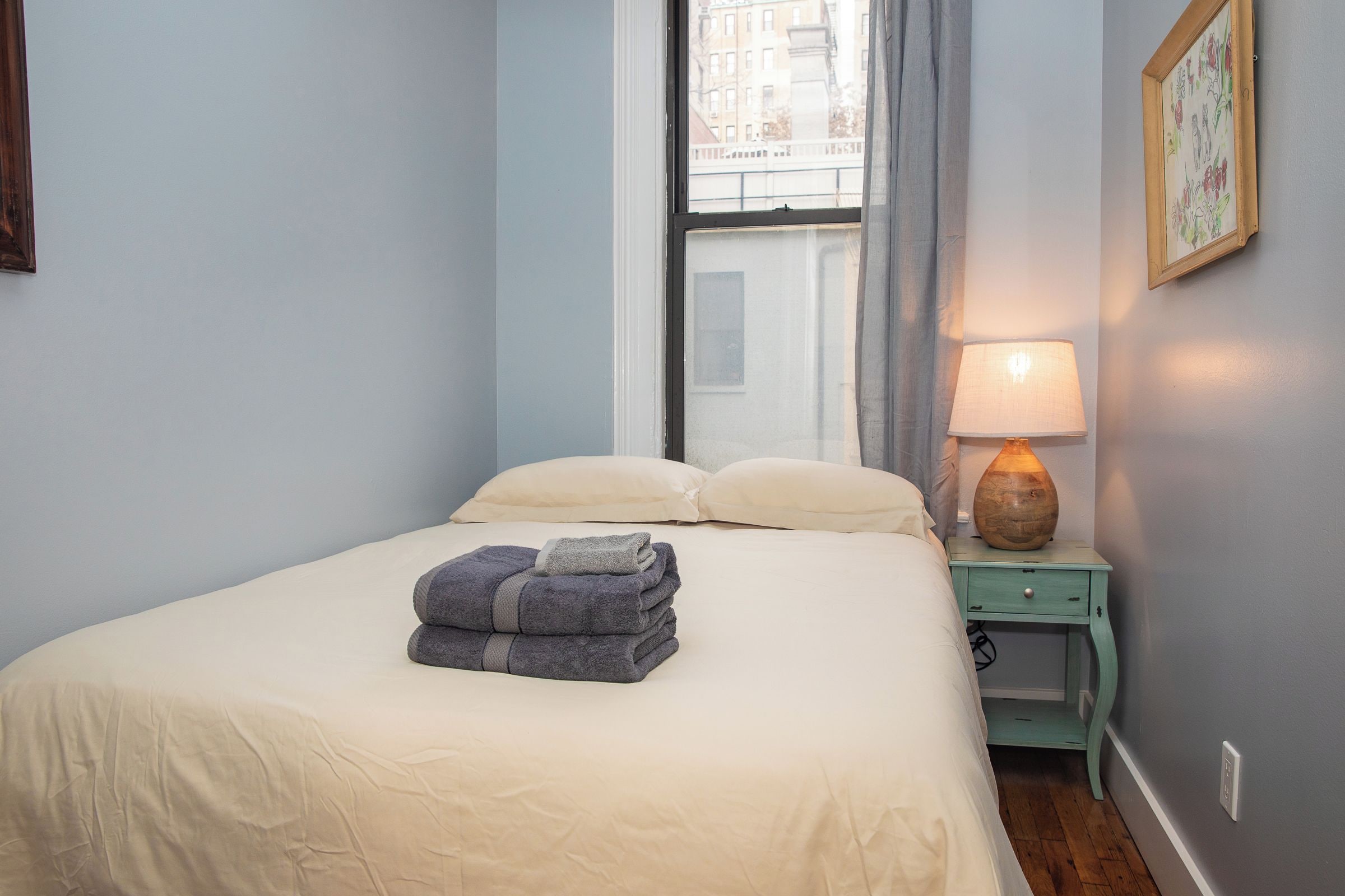 A neatly made bed with a folded towel, a bedside lamp, and a window with curtains.