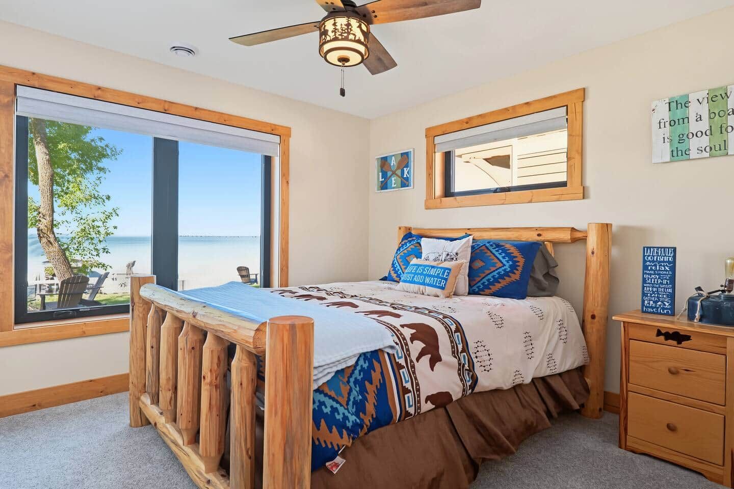 A bedroom with a large window, a bed with patterned bedding, a wooden headboard, a nightstand, and a ceiling fan.