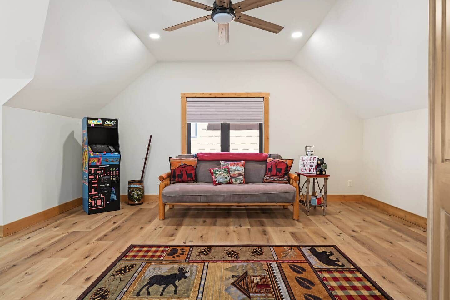 Ein Schlafzimmer im Loft-Stil mit schrägem Dach, Holzboden, einer Couch, einem kleinen Tisch, einem Fenster und einem Deckenventilator.