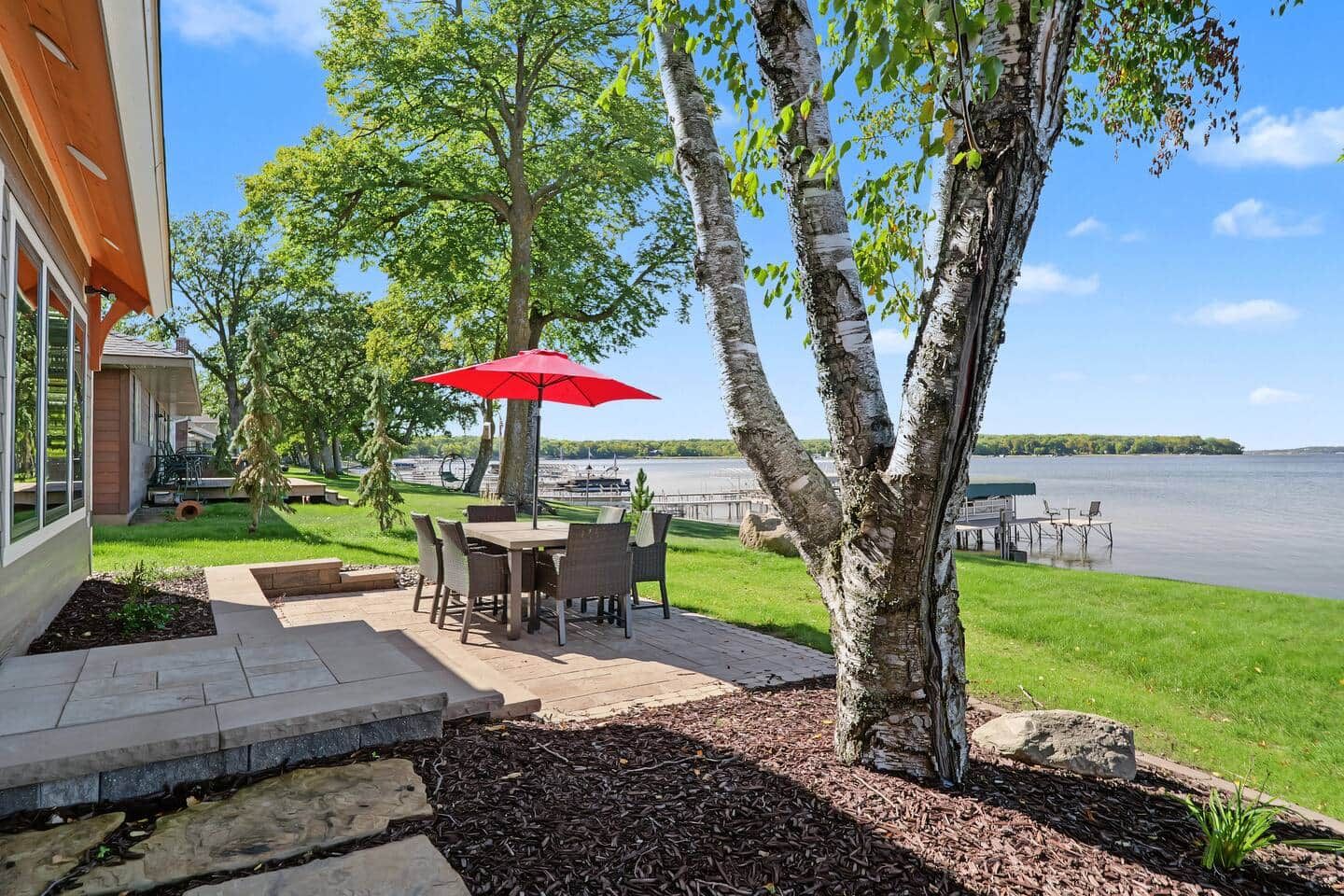 A patio with outdoor furniture and a red umbrella, overlooking a lake.