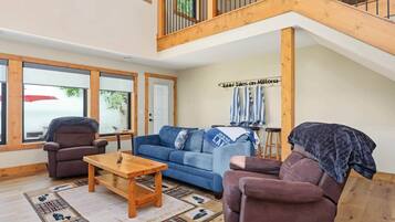 A spacious living area with a high ceiling, wooden beams, a blue sofa, two armchairs, a wooden coffee table, and a patterned rug.
