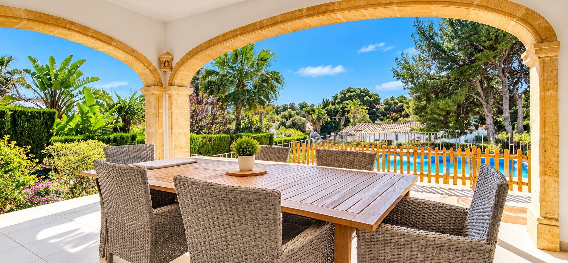 A covered outdoor dining area with a wooden table and wicker chairs, overlooking a pool and lush greenery.