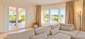 A modern living room with a sofa, a dining table with chairs, and a view of a garden through large windows. - Villa 'Vera' with sea view, Wi-Fi, heated pool and air conditioning (Balcó al Mar)
