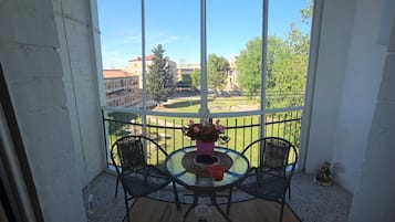 A covered balcony with two chairs and a table, a potted plant, and a view of buildings and trees.