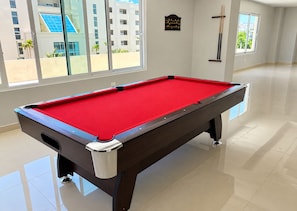 A pool table in a room with large windows, a clock on the wall, and a view of buildings outside. - Apartment with an ocean view (Playa Juan Dolio)