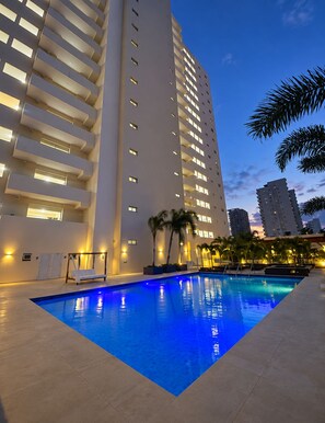 A swimming pool with a modern design, surrounded by a building with multiple floors and balconies, and palm trees. - Apartment with an ocean view (Playa Juan Dolio)