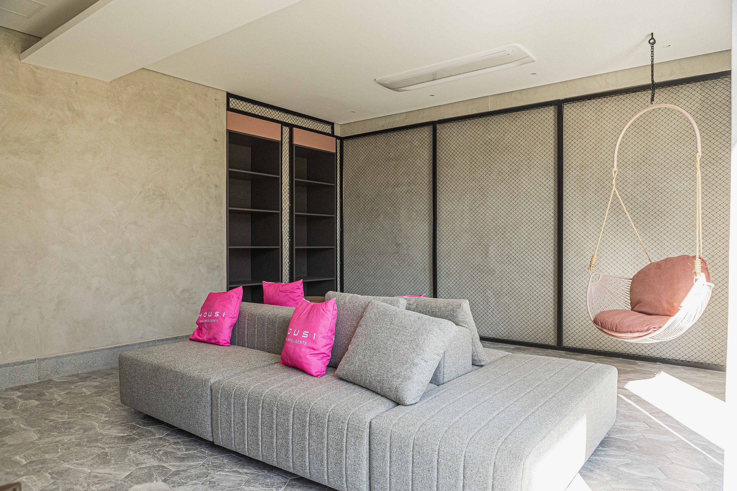 Un salon moderne avec un canapé gris, des coussins roses, un fauteuil suspendu et une grande fenêtre.