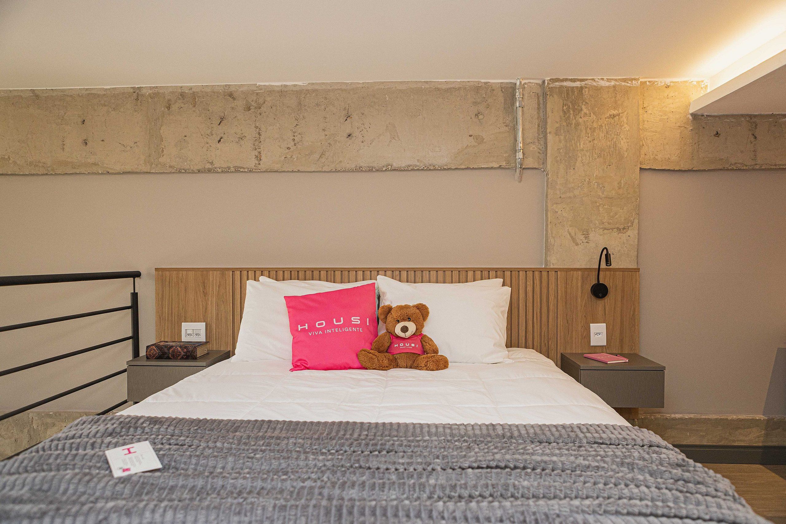 A hotel room with a bed, a bedside table, a nightstand, a teddy bear, and a wall-mounted light fixture.
