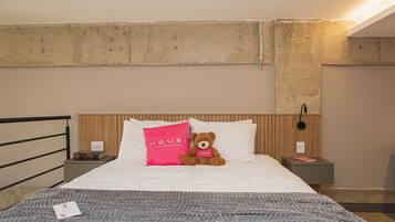 A hotel room with a bed, a bedside table, a nightstand, a teddy bear, and a wall-mounted light fixture.
