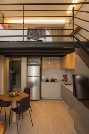 A compact kitchen with a built-in refrigerator, a dining area with a table and chairs, and a staircase leading to an upper sleeping area. - Housi Delphos (Belo Horizonte)