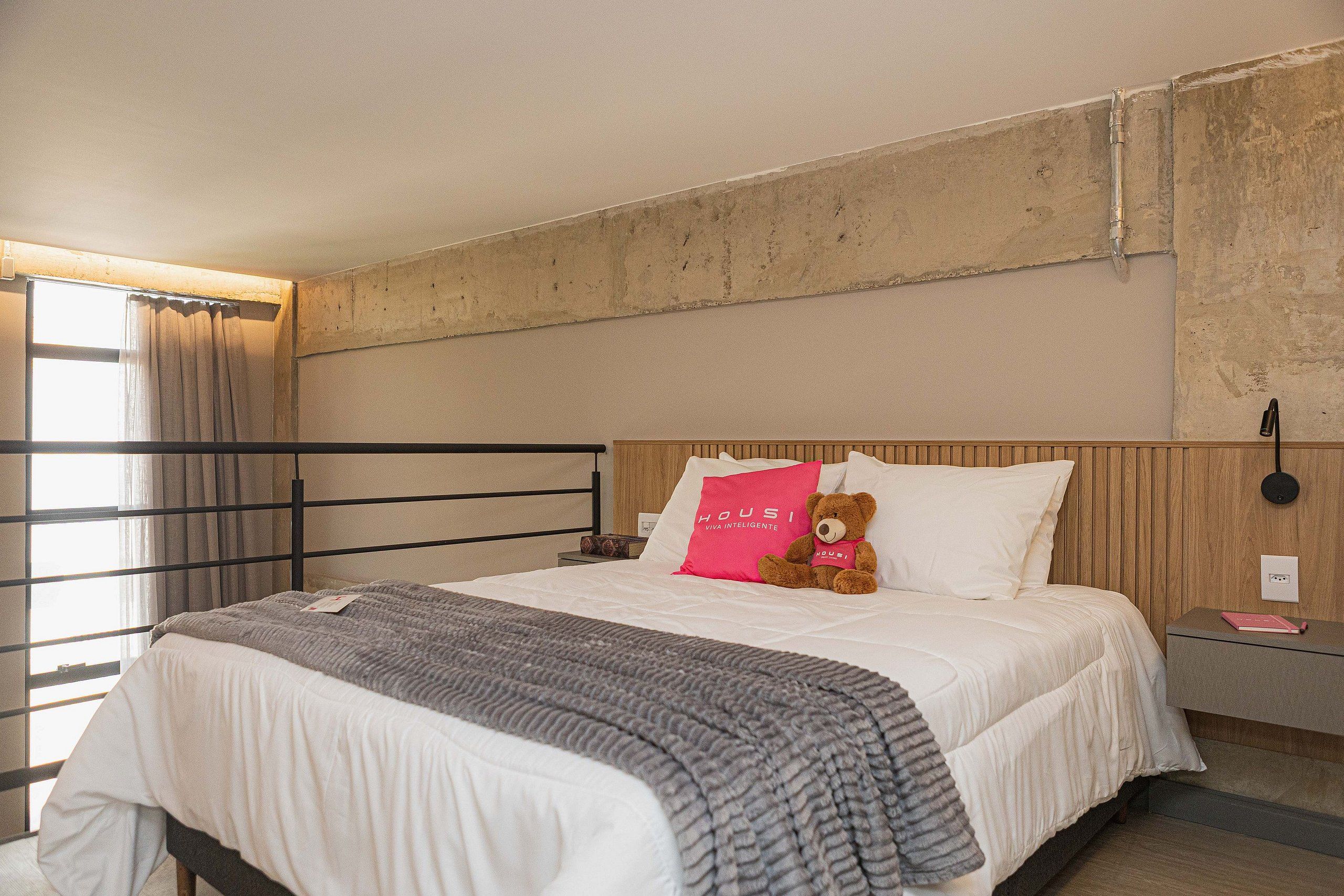 Una habitación de hotel con una cama, una mesita de noche, un oso de peluche y una ventana con cortinas.