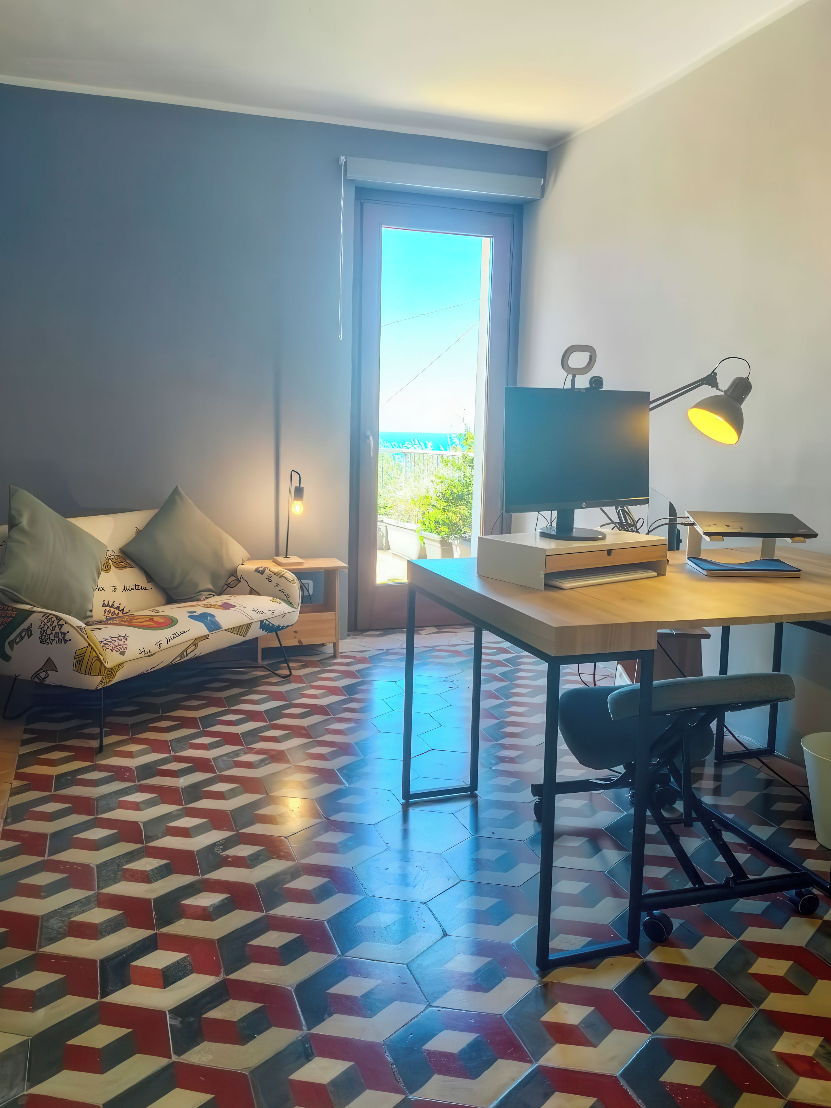 A modern room with a desk, chair, and a sofa with cushions. There is a window with a view of the outdoors.