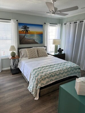 A bedroom with a bed, a ceiling fan, a painting of a beach scene, a nightstand with a lamp, and a window with curtains. - Beautiful 2-bedroom, 3 bed house on canal in Saint James City. (Saint James City)