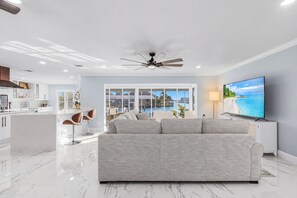 A modern living room with a large flat-screen TV, a grey sofa, and a view of a pool and beach through large windows. - Panoramic Water Views • Gulf Access • No Bridges (Cape Coral)